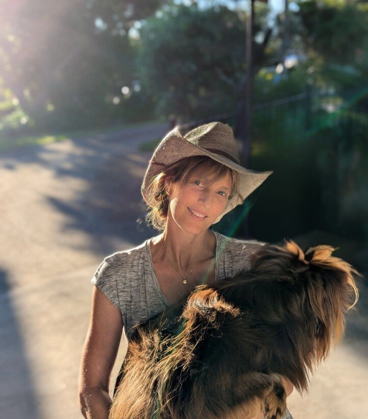 A warm, sunlit portrait of a woman with a gentle smile, wearing a grey t-shirt and a wide-brimmed woven straw hat. She is holding Monte, a brown Miniature American Shepherd with thick, fluffy fur, close to her chest. Sunlight streams in from the upper left, creating a soft glow and a slight lens flare. The background shows a shaded outdoor path with trees