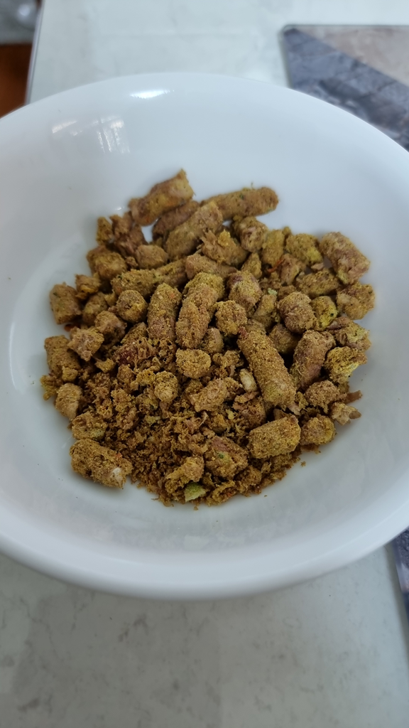 Here is the metadata for the dry Frontier Pet food image, written in Australian English for your blog. Alternative Text "A close-up high-angle shot of a white ceramic bowl containing dry, freeze-dried pet food. The food consists of various-sized cylindrical nuggets and crumbles in an earthy brown-gold colour with visible flecks of green herbs. The bowl is set on a light-coloured stone-textured kitchen benchtop