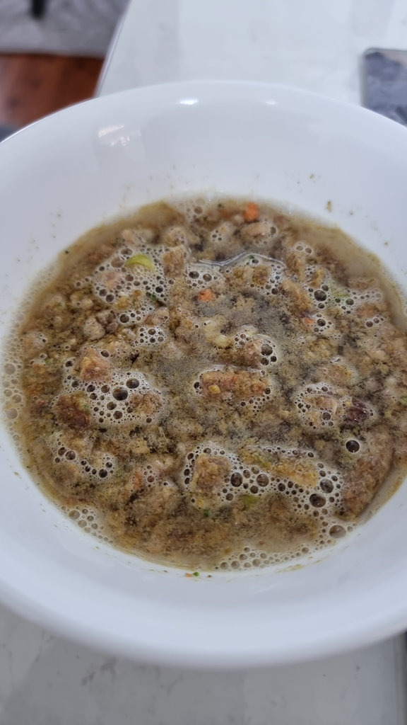 A close-up, high-angle shot of a white ceramic bowl containing rehydrated pet food. The food consists of small, textured brown nuggets and visible orange vegetable pieces mixed into a dark, bubbly broth. The bowl is set on a light-coloured kitchen benchtop with a blurred wooden floor in the background.