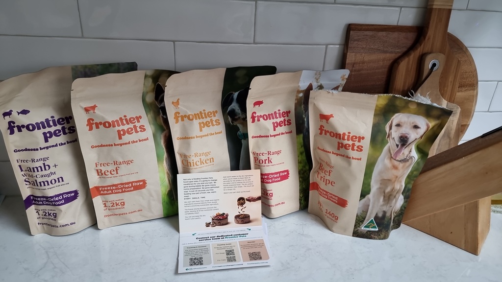 Here is the metadata for the Frontier Pets product range, written in Australian English for your blog. Alternative Text "A wide-angle horizontal shot of five different Frontier Pets dog food and treat bags lined up on a white marble-patterned kitchen benchtop. The bags feature varieties such as Free-Range Lamb + Wild-Caught Salmon, Free-Range Beef, Free-Range Chicken, Free-Range Pork, and Free-Range Beef Tripe. A small instructional pamphlet with QR codes sits in the foreground, and a wooden cutting board and knife block are visible to the right against a white tiled splashback.