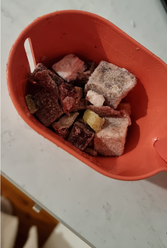 Here is the metadata for Monte’s frozen raw food portion, prepared in Australian English for your blog. Alternative Text "A top-down close-up shot of an orange plastic measuring scoop filled with frozen raw dog food cubes. The cubes vary in colour, including dark red, pale pink, and greyish-brown, with some showing a light dusting of frost. A single small, bright yellow-green vegetable piece is visible among the protein cubes. The scoop is held over a white stone-textured benchtop.