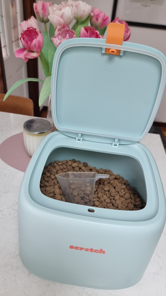 A vertical eye-level shot of a modern, light teal pet food storage container sitting on a white stone kitchen benchtop. The container has an open lid and the brand name 'Scratch' printed in orange on the front. Inside, the box is filled with brown, disc-shaped kibble, with a clear plastic measuring cup resting on top. In the background, a vase of pink and white tulips adds a soft, decorative touch to the scene.
