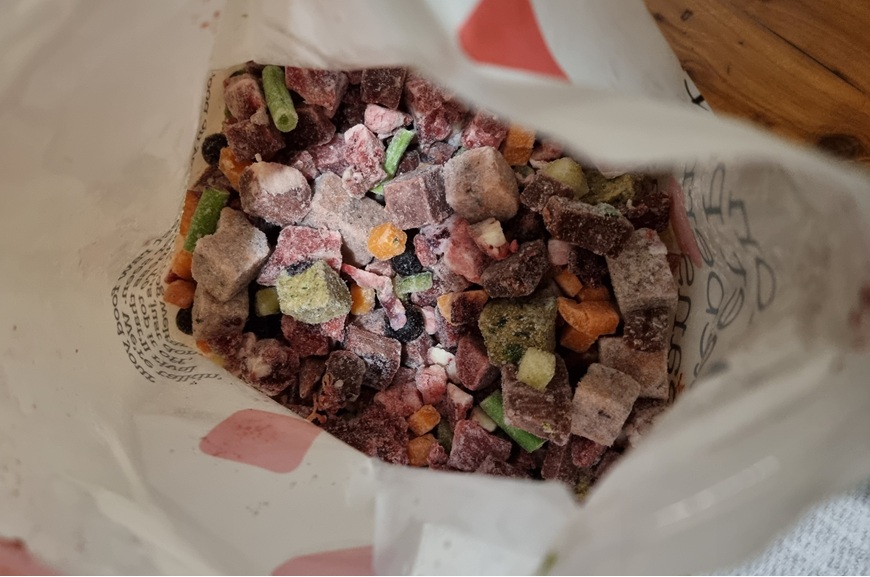 A top-down close-up shot looking into an open white bag filled with frozen raw pet food. The contents consist of small, colourful frozen cubes in shades of deep red, purple, and greyish-brown, mixed with bright green bean segments and small orange vegetable cubes. The interior of the bag shows printed black text.
