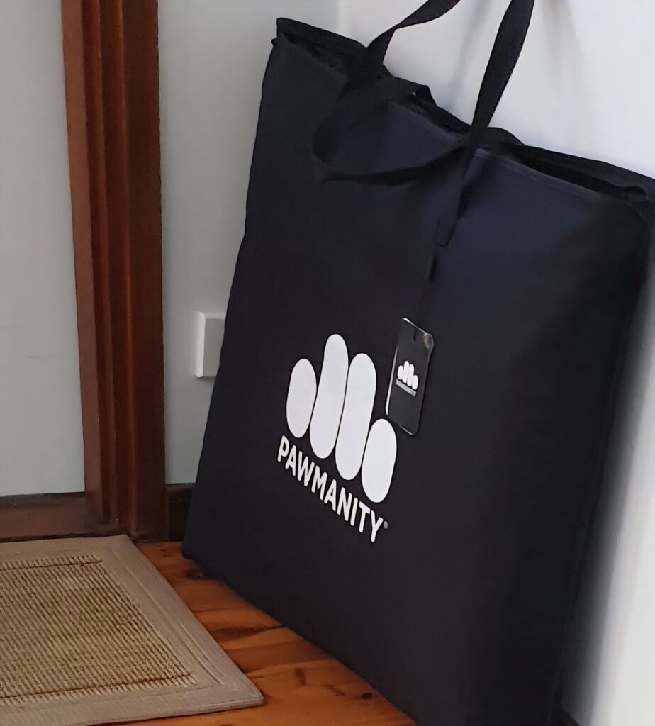 A large black storage bag with the white Pawmanity logo and "PAWMANITY" text printed on the front. The bag is leaning against a white wall next to a wooden door frame and a woven mat on a timber floor.