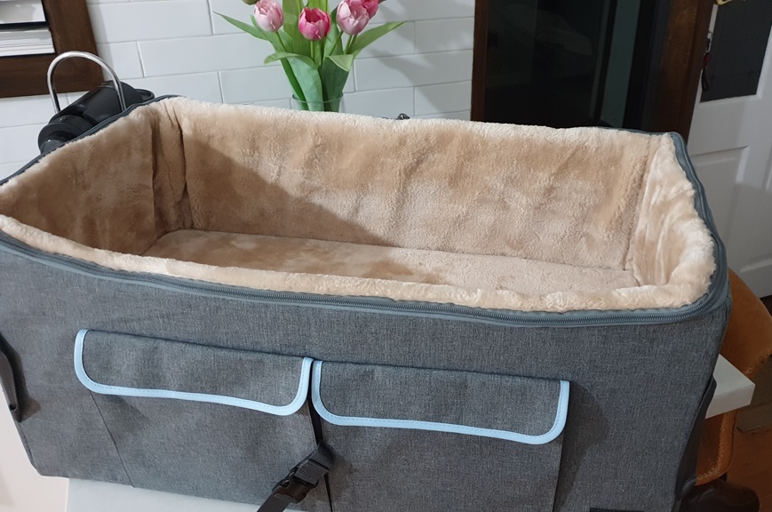 A close-up, high-angle shot of a brand-new Modern Pets elevated dog car seat sitting on a white table indoors. The seat features a durable grey fabric exterior with two front pockets trimmed in light blue. The interior is lined with a thick, plush, cream-coloured faux-fur material that looks incredibly soft. In the background, a vase of bright pink tulips and a white tiled wall add a touch of home styling.