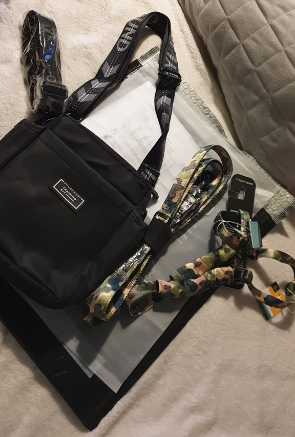 A flat-lay photo on a white textured blanket shows a black Stylish Hound OG Pouch 3.0 with an adjustable patterned strap. Beside it are a matching camouflage-patterned lead and a training long line, still with their tags on, resting on top of a grey shipping mailer. A grey quilted pillow is visible in the corner.