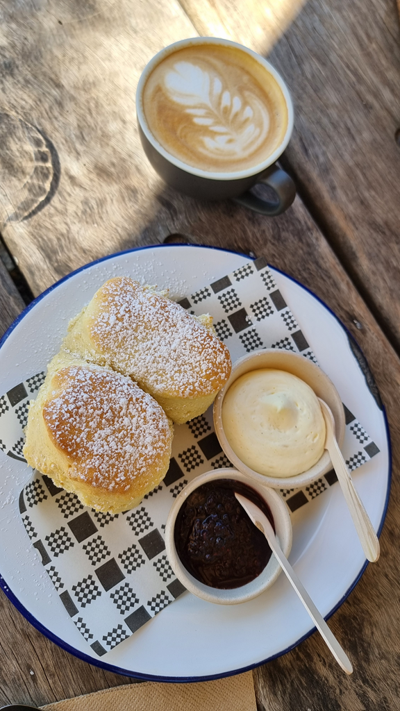 A vertical high-angle shot of two large, golden-brown scones dusted with icing sugar, served on a white plate with a blue rim. The scones sit on a black and white checkered paper napkin alongside two small white bowls containing thick cream and dark berry jam. A grey ceramic cup of coffee with latte art is visible in the background on a weathered timber table.
