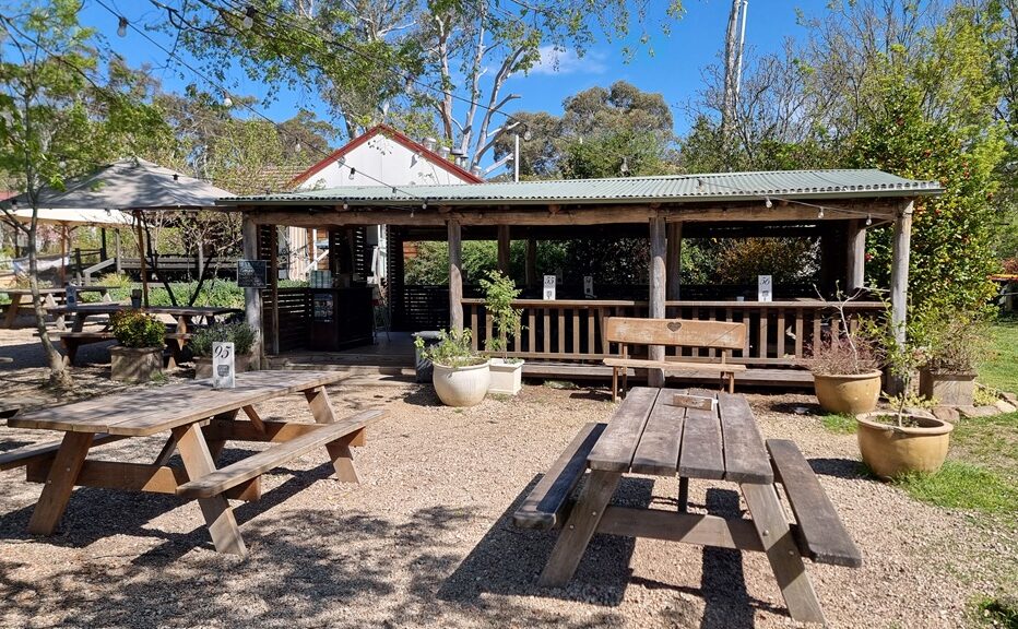 A horizontal wide-angle shot of an outdoor garden cafe with several timber picnic tables set on a gravel surface. A rustic timber pavilion with a corrugated iron roof provides a covered seating area in the background, surrounded by green trees and potted plants.