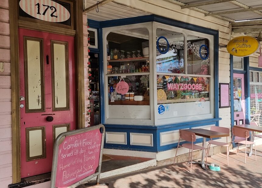 A horizontal eye-level shot of a charming café storefront with a vintage aesthetic. The exterior features soft pink weatherboard walls and a bright pink door with cream panels. A large bay window with dark blue trim displays a 'Wayzgoose Diner' sign and various cafe items. Outside, small timber tables and pink metal chairs sit on the brick pavement, with a teal pet water bowl visible on the ground. A pink chalkboard sign advertises 'Comfort Food' and 'Flowerpot Scones'