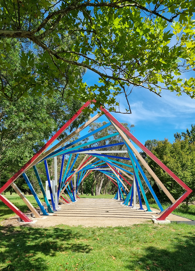 A vertical eye-level shot of a modern outdoor art installation consisting of a series of large, colourful diamond-shaped metal frames. The frames are painted in alternating shades of red, blue, turquoise, and tan, arranged in a row to create a tunnel-like perspective on a gravel path. The installation is set on a lush green lawn surrounded by established trees under a bright blue sky with wispy clouds.