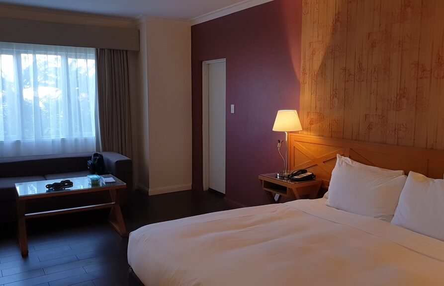 An interior shot of a hotel room with dark wood flooring and a purple accent wall. A large bed with white linens is in the foreground, next to a wooden nightstand with a lamp. In the background, a dark grey sofa sits beneath a window with sheer white curtains. A glass-topped coffee table holds a few personal items.
