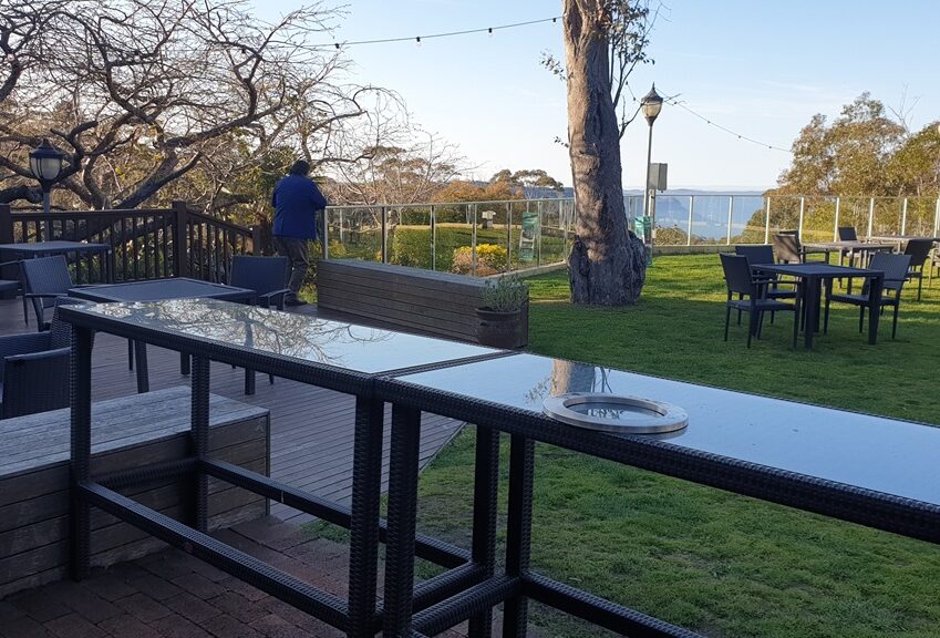 An outdoor hotel terrace featuring a wooden deck and a green lawn area with several black wicker tables and chairs. In the foreground, a long, glass-topped bar-height table sits on a brick patio. A large tree and a glass safety railing overlook a distant mountain valley under a clear blue sky