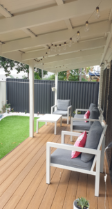 A covered wooden deck with light timber planks features a white outdoor lounge set with grey cushions and bright pink accent pillows. A string of warm Edison-style bulbs hangs from the white pergola rafters. Beyond the deck is a neat green synthetic lawn bordered by white pebbles and a dark charcoal grey fence.