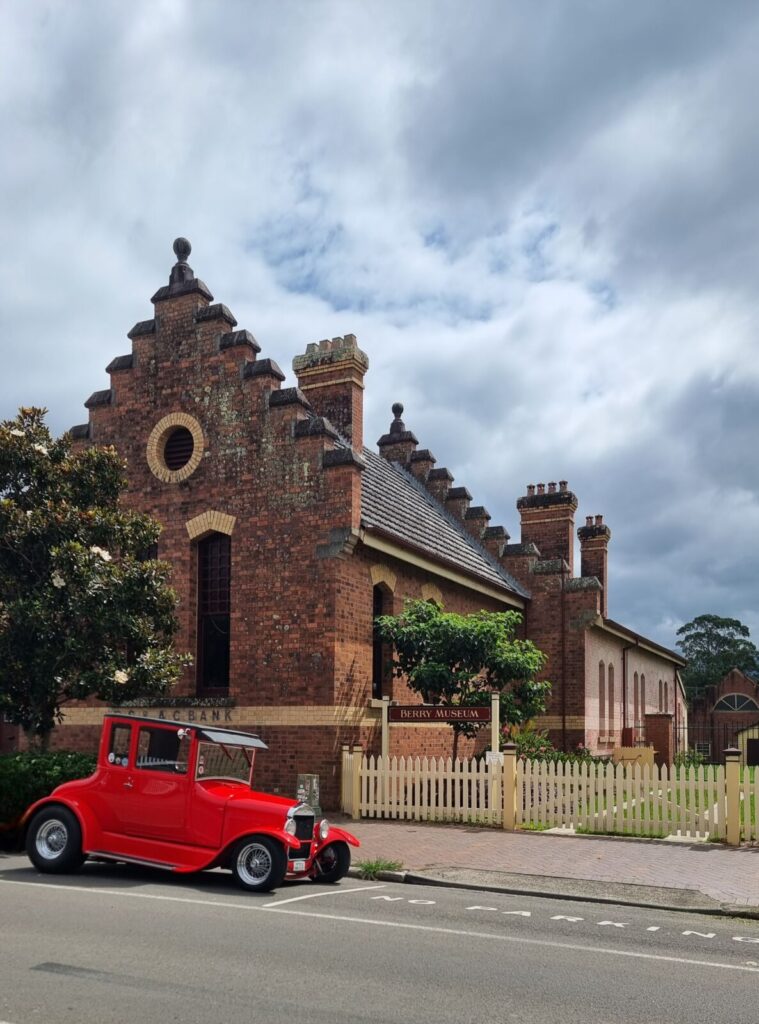 A bright red vintage hot rod car parked on a street in front of the historic Berry Museum. The museum is a grand, red-brick building with ornate gables and chimneys, situated behind a white picket fence under a dramatic, cloudy sky