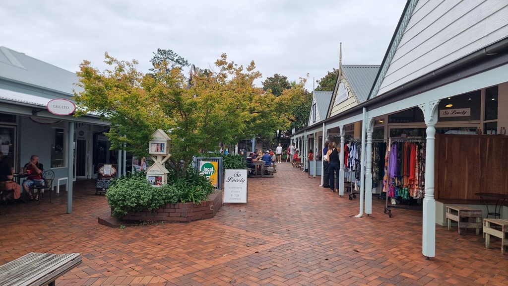 A wide-angle photograph of an outdoor brick-paved shopping courtyard in Berry. On the right, white boutique storefronts with decorative pillars display colorful clothing racks. In the center, a small tree with autumn leaves stands near a white sign that reads 'So Lovely,' and people can be seen sitting at outdoor tables in the background.