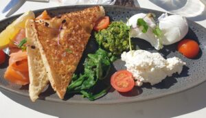 A top-down view of a gourmet breakfast plate on a white outdoor table. The dark speckled oval plate contains two poached eggs, two slices of thick toasted sourdough, a pile of smoked salmon with a lemon wedge, a scoop of green pesto, a portion of white ricotta cheese, and fresh wilted spinach, garnished with halved cherry tomatoes and microgreens.