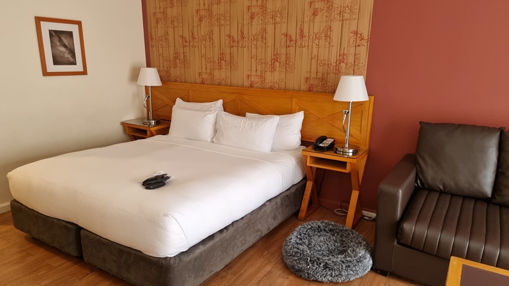 A bright hotel room interior featuring a large bed with white linens and a wooden headboard against a red accent wall. Two modern bedside lamps with white shades sit on wooden nightstands. A grey fluffy round pet bed is on the light timber floor next to a dark brown leather sofa. A framed black-and-white art piece hangs on the cream-colored wall.