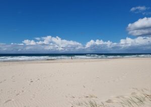 A wide-angle landscape of Broadbeach on a sunny day. In the foreground, a vast expanse of pale, rippled sand is marked with scattered footprints and sparse beach grass. The deep blue ocean has small white breaking waves, under a bright blue sky filled with a low horizontal line of fluffy white clouds.
