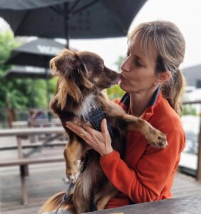A heartwarming close-up portrait of Caroline holding Monte at an outdoor cafe table at Bisou Bakery. Caroline, with light brown hair and a bright orange fleece, is leaning in to kiss Monte’s nose. Monte, a mahogany Miniature American Shepherd in a black harness, looks at her affectionately. The background shows blurred dark patio umbrellas and wooden tables.