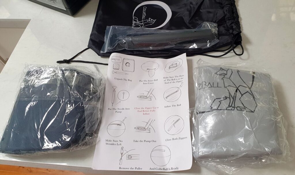 A flat-lay photo of the contents of the CollieBall drawstring bag, unboxed on a white marble-style countertop. In the center is a printed instruction sheet titled "CollieBall Is Ready" with diagrammed steps for assembly. To the left, a blue object, possibly the ball cover, is wrapped in clear plastic. Above it is a long, narrow pump with a nozzle. To the right is another grey object in a plastic bag with a geometric dog logo and the text "BALL". The black CollieBall drawstring bag is in the top-right.