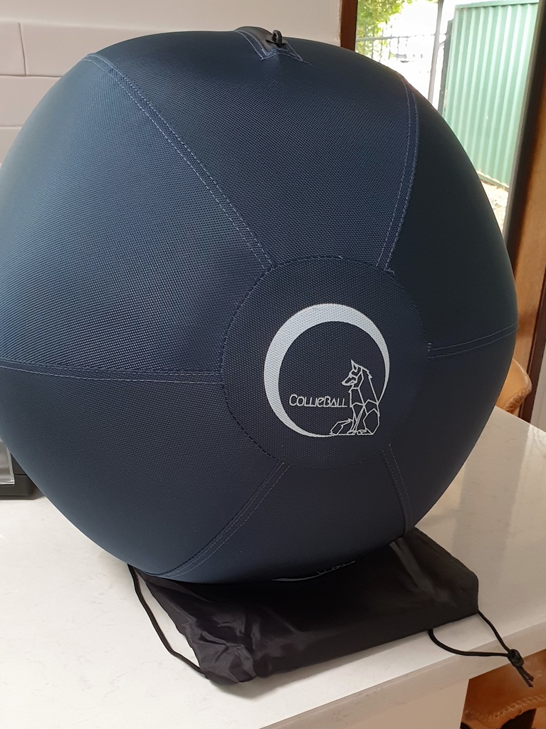 A large, fully inflated navy blue CollieBall sits on a white kitchen benchtop. The durable fabric cover is taut, showing the white geometric dog logo clearly in the centre. The black drawstring storage bag lies flat underneath the ball.