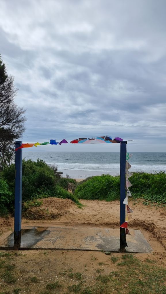 A vertical outdoor photograph of a beach entrance at Culburra Beach. Two blue wooden posts form a gateway, draped with a colorful string of bunting flags fluttering in the wind. The gateway frames a view of a sandy path leading down to the ocean under a cloudy sky.
