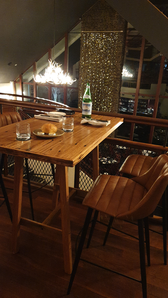 A vertical indoor shot taken at night showing a high-set timber dining table and two tan leather bar stools positioned on a mezzanine level overlooking a hotel lobby. The table is set for two with glass tumblers, white napkins, and a small plate of bread rolls, with a large bottle of sparkling water in the centre. An antler-style chandelier hangs above, and a tall stone pillar decorated with fairy lights is visible through a large glass window in the background.