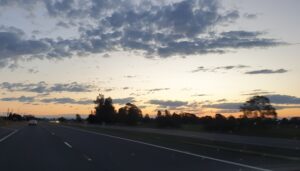 A view from a car dashboard looking onto a multi-lane highway at dawn. A silver car is visible in the distance on the left. The horizon glows with a soft orange and yellow sunrise, while the upper sky is filled with dark, scattered clouds. Silhouetted trees and utility poles line the side of the road.