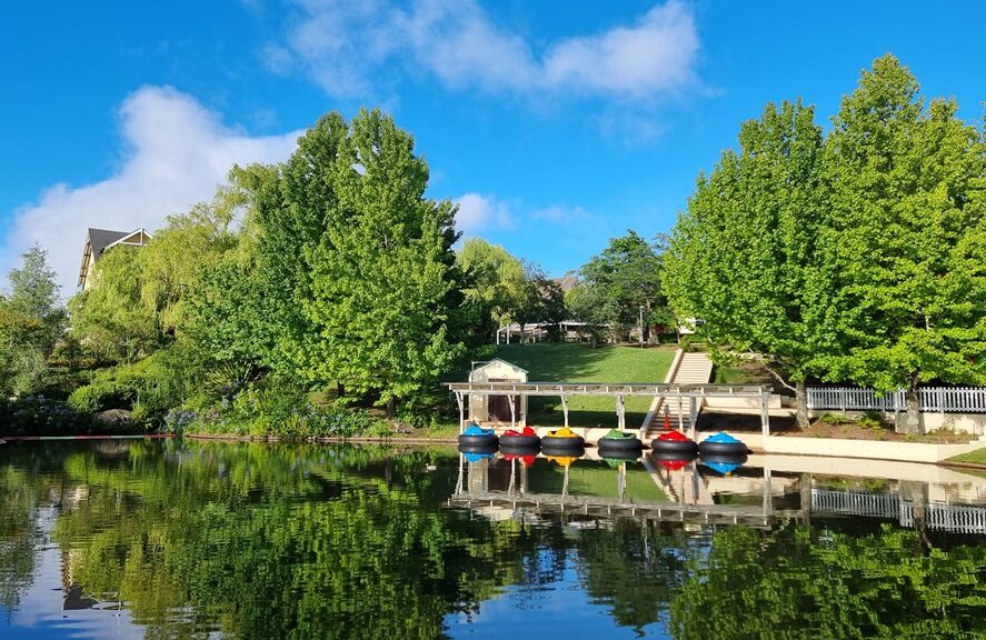 A bright, wide-angle landscape of a hotel's recreation area featuring a large, still pond that perfectly reflects a vibrant blue sky with soft white clouds. A row of colorful bumper boats in red, blue, green, and yellow are docked under a white-roofed pavilion along the water's edge. Lush green grass slopes up to a set of stairs and a dense canopy of tall, bright green trees, with the gabled roof of a cream-colored building visible in the background