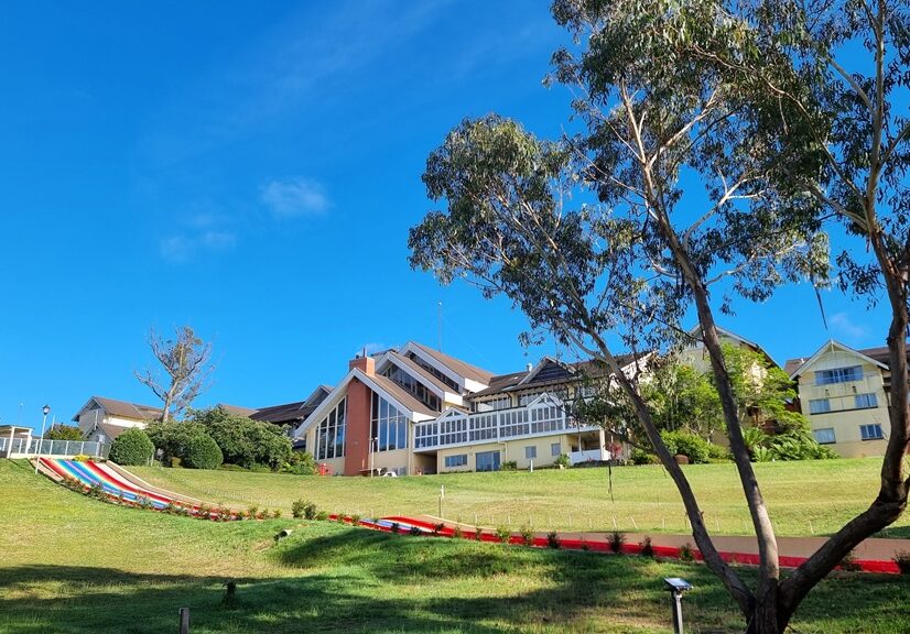 A wide-angle landscape shot of the Fairmont Resort building from the back lawn. A large, cream-colored hotel with multiple gabled roofs and a distinctive glass-walled section sits at the top of a bright green, sloping hill. To the left, a long, colorful rainbow-striped slide runs down the grass. A large, multi-trunked eucalyptus tree frames the right side of the shot under a clear blue sky.