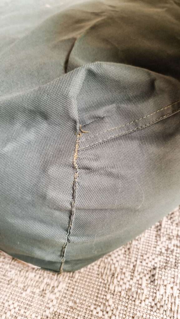 A close-up of a second repaired corner on a green canvas dog bed, showing dark, thick reinforcement stitching over a previous tear. The bed sits on a neutral-toned woven rug.