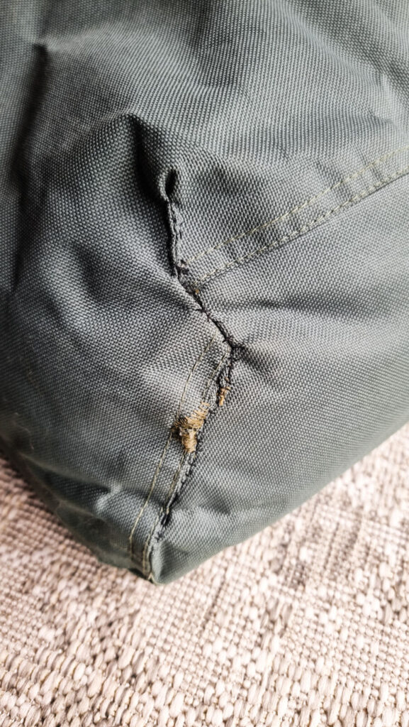 A macro close-up of a repaired seam on a green canvas dog bed. The heavy-duty fabric shows a vertical line of tight, slightly frayed stitching where a tear has been mended, resting against a textured cream rug.