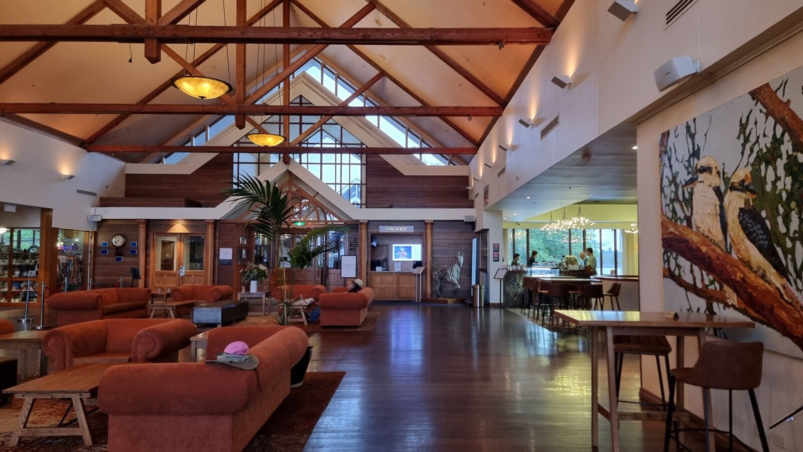 A wide-angle interior shot of a grand hotel lobby featuring high vaulted ceilings with prominent wooden beams and warm pendant lighting. The room has polished dark wood floors and several burnt-orange plush sofas arranged in seating clusters. On the right, a large painting of two kookaburras is displayed near a high wooden table with bar stools, leading into a brightly lit bar area.