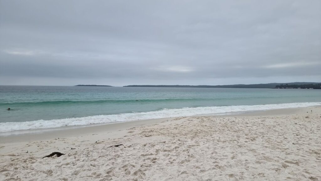 A wide landscape photograph of Hyams Beach under a heavy, overcast grey sky. The scene features bright white sand in the foreground, transitioning to turquoise and deep blue ocean water with gentle white waves breaking on the shore. A dark headland is visible on the horizon.