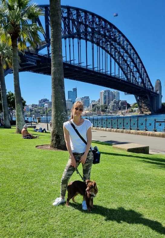 A woman with sunglasses and a white t-shirt stands on a bright green lawn in front of the Sydney Harbour Bridge. She is wearing camouflage-patterned pants and has the black Stylish Hound OG Pouch 3.0 slung across her body. Monte, the brown Miniature American Shepherd, stands patiently on a lead beside her under a clear blue sky.