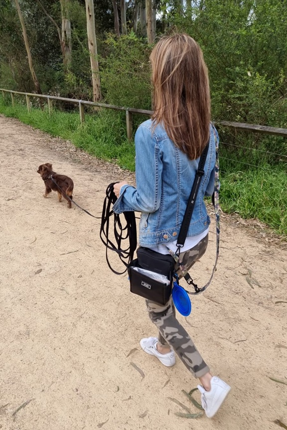 A throwback photo from behind of a woman with long hair, wearing a denim jacket and camouflage-patterned leggings, walking a young Monte on a sandy path. She is wearing the black Stylish Hound OG Pouch 3.0, which has a blue collapsible bowl clipped to it. In one hand, she manages a long black training line as Monte, a small brown Miniature American Shepherd puppy, walks ahead on a lead.