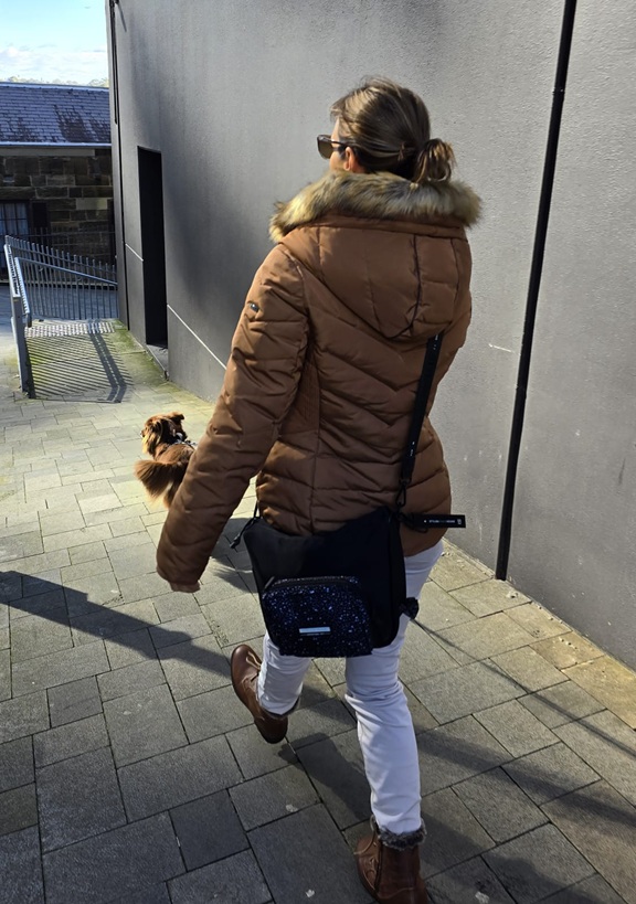 A woman with a ponytail, wearing a brown quilted puffer jacket and white trousers, walks away from the camera on a paved path. She is wearing a large black Stylish Hound Treat Pouch slung across her body, which features a smaller patterned pocket on the front. Monte, the brown Miniature American Shepherd, is walking just ahead of her.