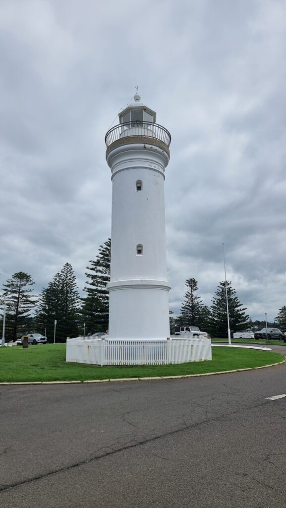 A vertical photograph of the Kiama Lighthouse, a tall, white cylindrical tower with a black railing around the lantern room at the top. The lighthouse is situated on a green lawn surrounded by a white picket fence, with tall pine trees and a cloudy sky in the background.