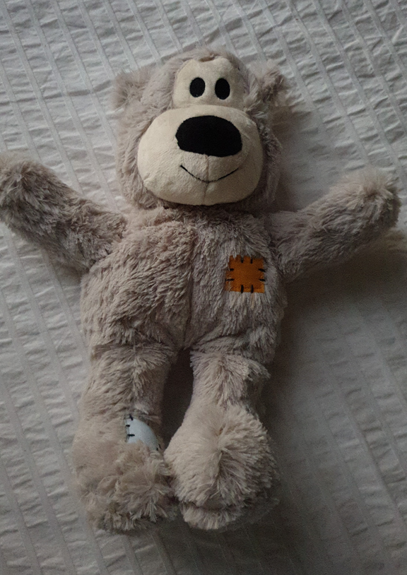 A top-down shot of a well-loved, light grey plush teddy bear lying on a white textured fabric surface. The bear has a round, cream-coloured snout with a large black nose and small black eyes. A small orange rectangular patch is stitched onto its chest, and a white patch is visible on its lower left leg. The plush fur appears soft and slightly matted from years of use.