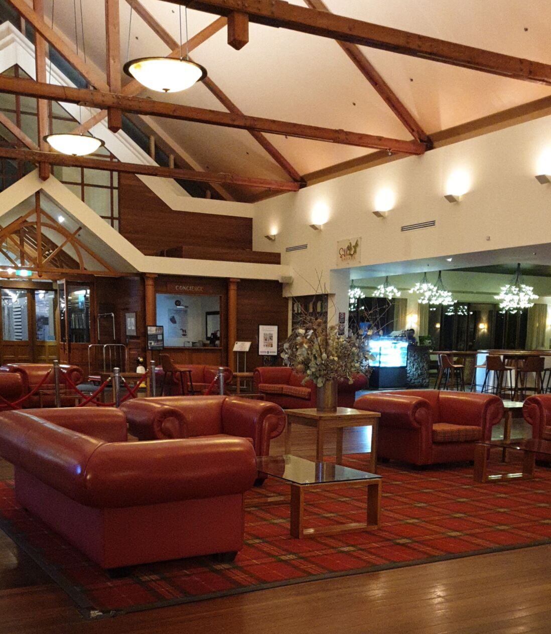 A spacious resort lobby with high vaulted ceilings and exposed timber trusses. The room features a warm wood-panelled concierge desk and several plush red leather sofas arranged on a red tartan rug. Large glass doors and a warm, glowing light from various chandeliers create a cozy, boutique atmosphere.
