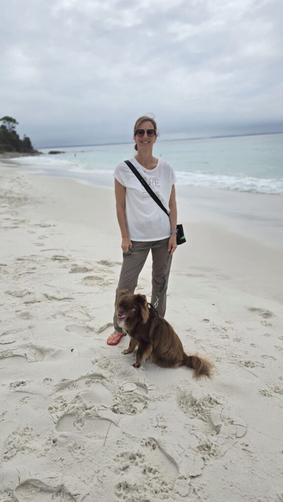 A full-length outdoor photograph of a woman, Caroline, standing on the white sand of Hyams Beach with her brown Miniature American Shepherd, Monte. Caroline is wearing a white t-shirt, olive green trousers, and sunglasses, holding Monte's leash. Monte is sitting obediently beside her on the sandy shore under an overcast sky.