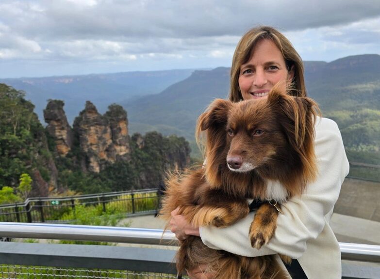 A horizontal medium shot of Caroline, a woman with shoulder-length brown hair wearing a white jacket, holding Monte, her mahogany Miniature American Shepherd, in her arms. They are standing at a lookout with a metal railing. In the background, the Three Sisters rock formation rises above the vast, green Jamison Valley under a cloudy sky