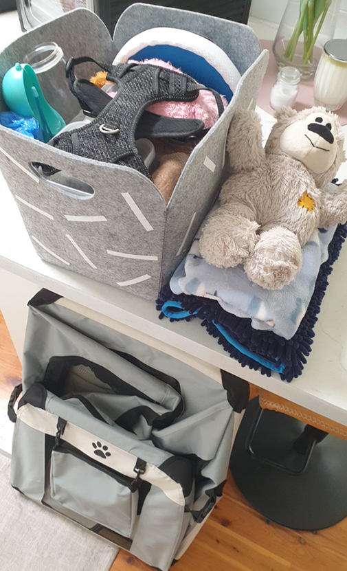 A high-angle shot of a white countertop organized with pet travel gear. A grey felt basket with a white geometric pattern is filled with a black harness, a blue portable water dispenser, and pink bedding. Next to it, a plush teddy bear sits on a folded blue patterned blanket and a dark navy microfibre mat. Below the counter, a grey and black soft-sided pet carrier with a paw print logo is visible on a light wood floor.