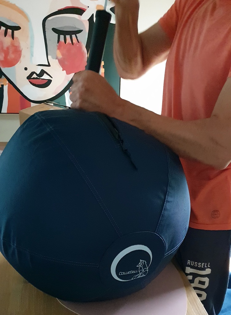 A close-up shot of the navy blue CollieBall being inflated on a timber table. A person in an orange shirt is using a black pump to finish the job. In the background, a modern "slow art" style painting of a face is visible on the wall.