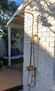 A sleek, wall-mounted outdoor shower with a brushed brass finish is installed on a white-painted brick wall. The unit features a large overhead rainfall showerhead, a handheld attachment, and hot and cold taps. It is positioned over a timber deck, with a glimpse of a green backyard and a black safety fence in the background.