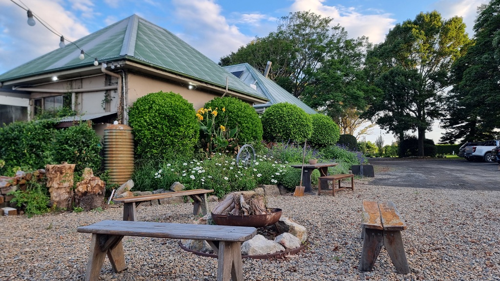 A wide-angle outdoor shot of the Butter Factory. The scene features a gravel courtyard with several rustic wooden benches surrounding a stone fire pit. A historic building with a green corrugated iron roof is in the background, surrounded by lush green hedges, white daisies, and yellow flowers under a blue sky with soft white clouds.