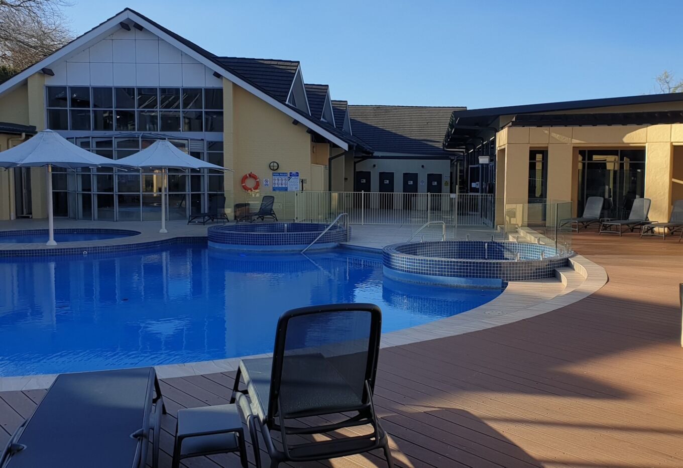 A large, bright blue outdoor swimming pool with two circular integrated spas sits at the center of a resort courtyard. The area is surrounded by a light timber deck with several dark grey lounge chairs. In the background, there is a large yellow building with multiple peaked rooflines and a glass-enclosed section under white umbrellas.