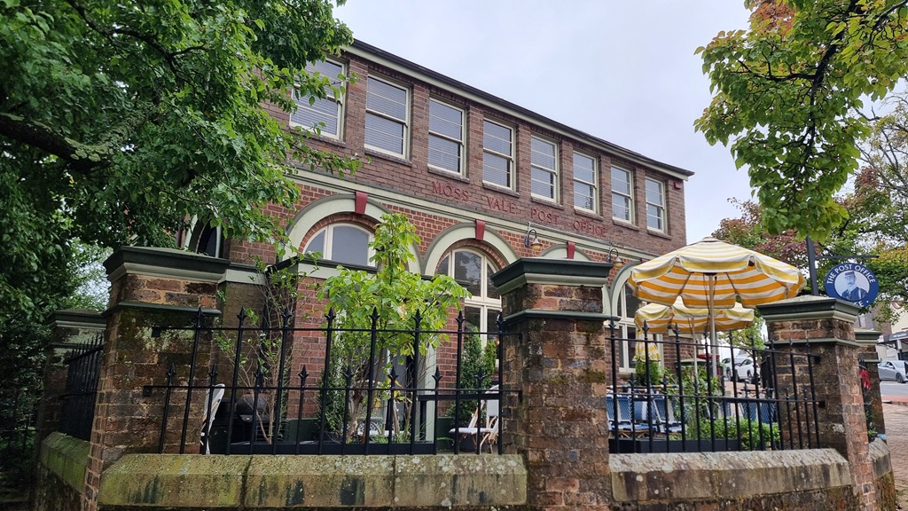 A historic two-story red-brick building labeled 'Moss Vale Post Office' now serving as a cafe. The building features large arched windows on the ground floor and a row of rectangular windows above. A black wrought-iron fence surrounds an outdoor patio with yellow-and-white striped umbrellas, partially shaded by lush green trees