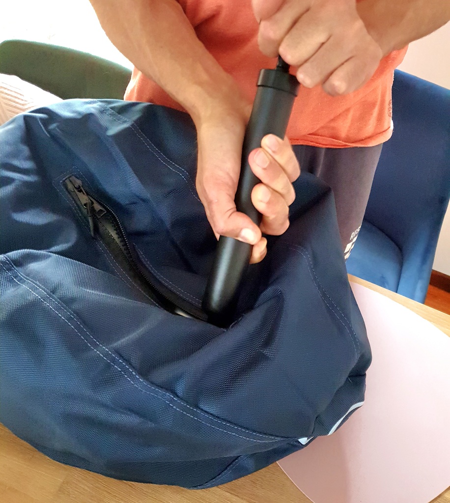 A close-up shot of hands using a black manual pump to inflate the inner ball through the zipped opening of the navy blue CollieBall fabric cover. The person is wearing an orange shirt, and the activity is taking place on a light timber tabletop.