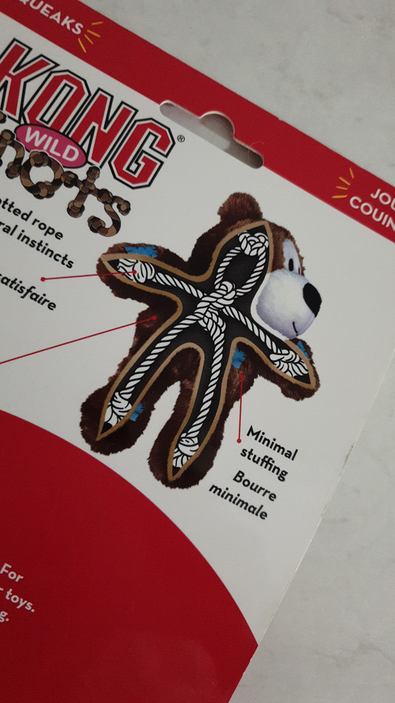 A close-up angled shot of the back of a KONG Wild Knots teddy bear package. The packaging shows a diagram of a brown plush bear with an internal skeleton made of sturdy, knotted white rope to illustrate durability. Red lines point to features like 'knotted rope' and 'minimal stuffing'. The background is a clean, white stone-textured benchtop.